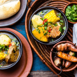 Steaming bowl of Latin-style chicken soup with vegetables, garnished with fresh cilantro and lime wedges, served in a traditional clay bowl.