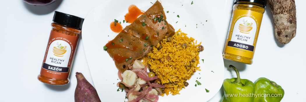 A delicious plate of Arroz con Gandules, the traditional Puerto Rican rice with pigeon peas, paired with a savory Puerto Rican pastel drizzled in red sauce, and a side of pickled root vegetables. The plate is complemented by Healthy Rican Sazón Seasoning and Adobo Seasoning jars, surrounded by fresh ingredients like sweet potatoes and green bell peppers, showcasing the best adobo and sazón seasonings for authentic Puerto Rican cooking.